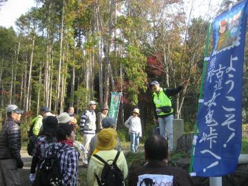 「ようこそ 古道女鬼峠へ！」と書かれたのぼり旗の横で、観光ボランティアガイドの男性が女鬼峠のコースの参加者に話をしている様子の写真